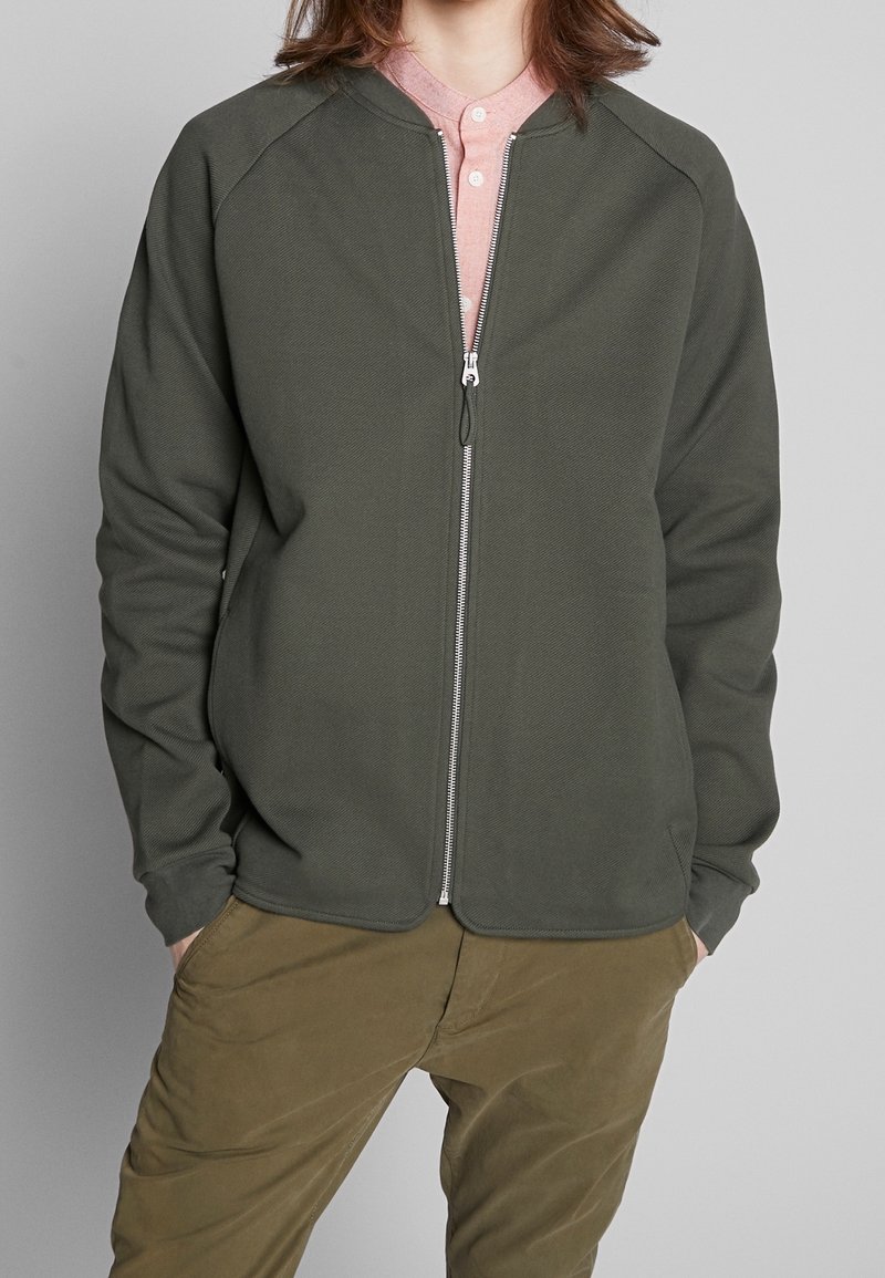 Personne portant une veste vert olive ouverte sur une chemise rose à col et un pantalon vert olive, les mains dans les poches, debout devant un fond gris.