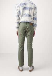 Henkilö, jolla on vihreät housut, valkoiset sukat, beiget liukupohjaiset kengät ja valkoinen pitkähihainen paita, jossa on sinisiä maisemakuvioita, seisoo selkä kameraan päin.