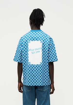 Άνδρας με dreadlocks γυρισμένος από πίσω, φοράει μπλε και άσπρο πουκάμισο καρό με το κείμενο «Carlo Colucci Est. 1978» στην πλάτη, συνδυασμένο με μπλε τζην.