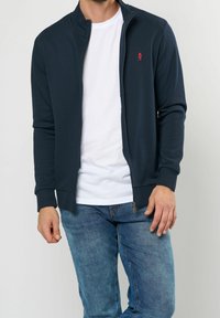 Mörkblå zip-up jacka med ribbade ärmar och krage; har en liten röd logotyp på bröstet och en slät tygstruktur. Bärs över en vit t-shirt.