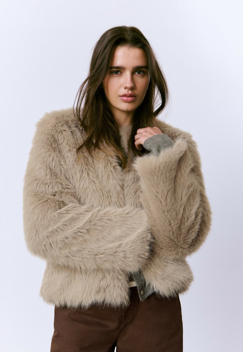 Mujer con cabello largo castaño que lleva una chaqueta de piel sintética beige sobre un suéter gris y pantalones marrones, posando con una mano cerca de su cara.
