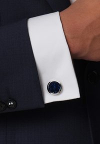 Navy suit with a textured fabric, featuring a white cuff and a silver cufflink with a round blue stone in the center.
