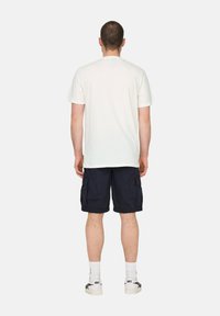 Hombre de pie de espaldas, vistiendo una camiseta blanca sencilla, pantalones cortos de carga azul marino, calcetines blancos y zapatillas Asics blancas, sobre un fondo blanco.