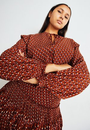 Γυναίκα με μακριά σκούρα μαλλιά φοράει ένα κόκκινο φλοράλ φόρεμα με μακριά μανίκια, στέκεται με τα χέρια σταυρωμένα, κοιτάζοντας με αυτοπεποίθηση στην κάμερα.