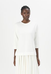 Maglione bianco, morbido e ampio, con maniche a tre quarti, scollo rotondo e un sottile logo dettagliato all'orlo. Tessuto morbido e design minimalista.