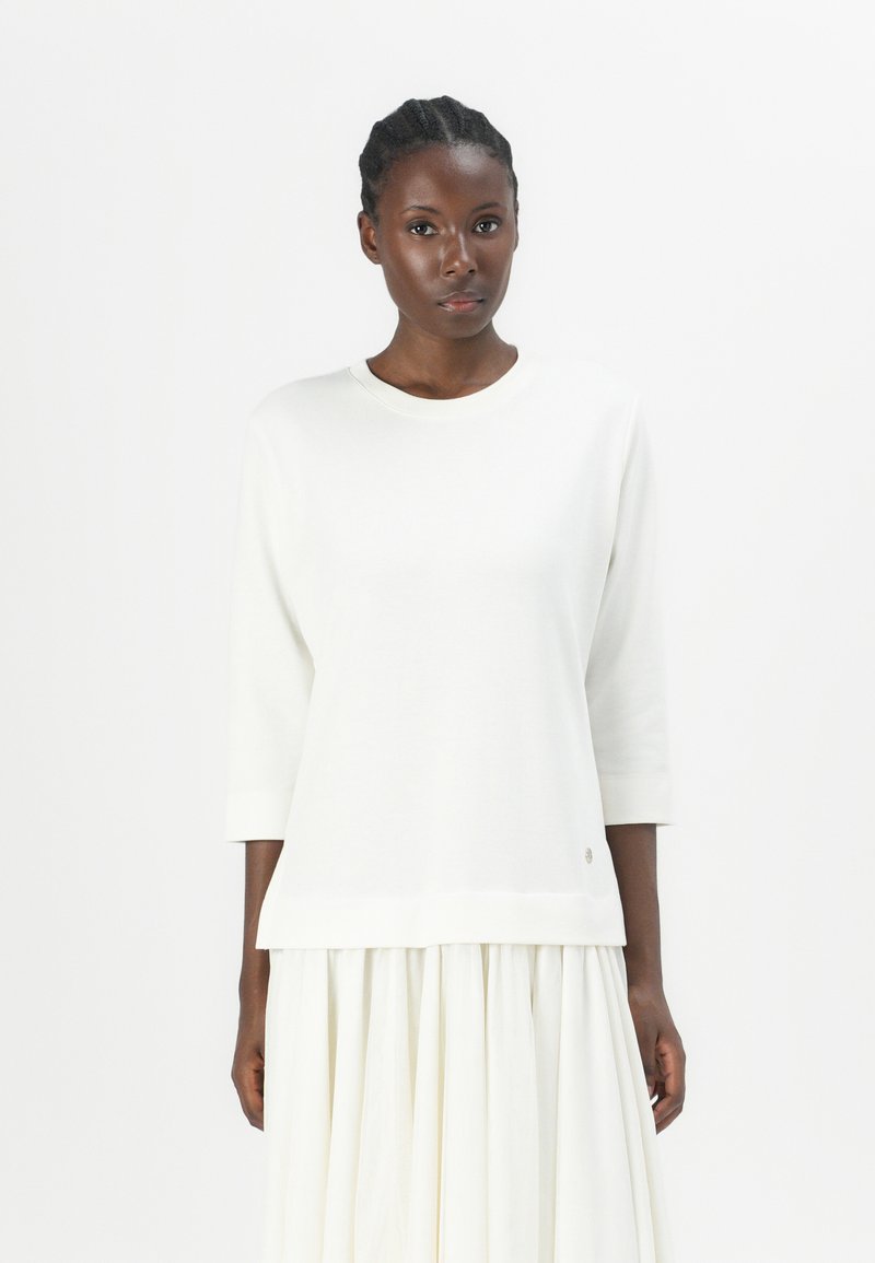 Maglione bianco, morbido e ampio, con maniche a tre quarti, scollo rotondo e un sottile logo dettagliato all'orlo. Tessuto morbido e design minimalista.