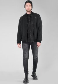 Giacca in pile nera con colletto alto, abbinata a una camicia nera con bottoni e jeans neri attillati. Berretto nero e stivali con suola in gomma.