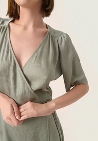 Zelené zavinovací šaty s texturovaným povrchem, krátkými rukávy a hlubokým výstřihem do V, zdobené detailním zapínáním na boku.