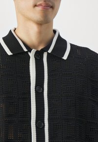 Homme portant une chemise en maille noire boutonnée avec une bordure blanche au col et sur la patte de boutonnage, montrant partiellement le visage et le cou.