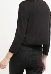 En person i en figursydd svart långärmad topp och högmidjade svarta jeans sedd bakifrån mot en enfärgad vit bakgrund.