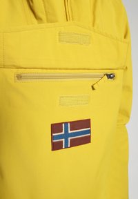Gele waterdichte jas met een ritssluiting aan het zakje en een gestikte Noorse vlagpatch. Soepele textuur met zichtbare stikdetails.