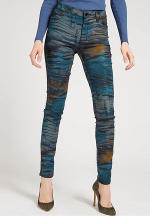 Slim-fit pants with blue, black, and brown abstract patterns worn with dark green high-heeled shoes and a blue ribbed long-sleeve top.