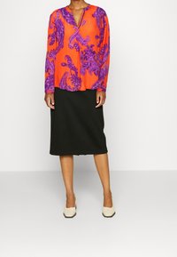 Blusa a maniche lunghe di un acceso arancione con motivo paisley viola, abbinata a una gonna nera al ginocchio. Tessuto morbido e vestibilità rilassata.