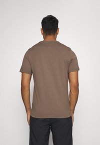 Hnědé tričko s krátkým rukávem a kulatým výstřihem, se hladkým a přiléhavým materiálem, z pohledu zezadu, v kombinaci s tmavými šortkami.