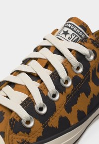 Κοντινό πλάνο σε αθλητικό παπούτσι Converse από καμβά με μοτίβο λεοπάρ, λευκά κορδόνια και μεταλλικά περβόλια, με το λογότυπο της μάρκας στη γλώσσα.