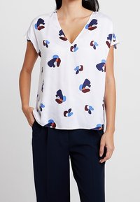 Blusa bianca a maniche corte con scollatura a V, caratterizzata da modelli astratti in blu, rosso e blu navy. Tessuto leggero con una texture liscia.