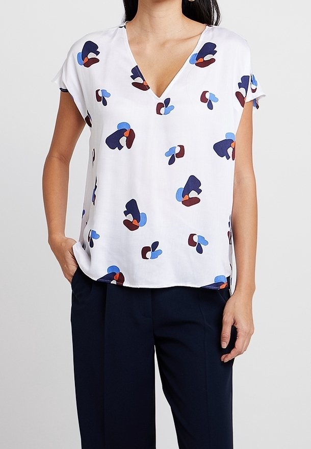 Blusa bianca a maniche corte con scollatura a V, caratterizzata da modelli astratti in blu, rosso e blu navy. Tessuto leggero con una texture liscia.