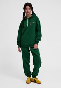 Zielona bluza z kapturem i pasujące joggery wykonane z miękkiego materiału, z kieszenią kangurką, sznurkami do ściągania i akcentem logo. Połączone z żółtymi sneakerami.