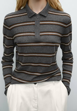 Vrouw draagt een grijs poloshirt met lange mouwen met bruine en witte horizontale strepen, in een witte broek met hoge taille gestopt.