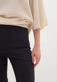 Crème, oversized gebreide trui met brede mouwen en een zachte textuur, gecombineerd met strakke zwarte broeken. Hand geplaatst langs de naad van de broek.