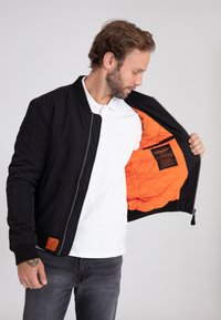 Zwarte bomberjack met elastische manchetten, voorzien van een gewatteerde oranje binnenkant en een label aan de binnenkant van de linkerkant. Model draagt een wit shirt.