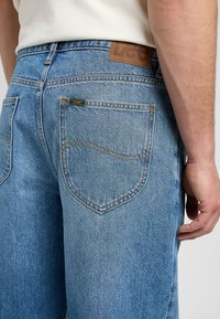 Homme portant un jean en denim bleu clair de la marque Lee et une chemise blanche, montrant une poche arrière et une main sur le côté.