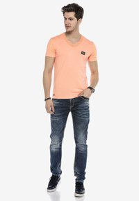 Kurzärmliges, V-Ausschnitt T-Shirt in leuchtendem Koralle mit Akzentnähten, kombiniert mit hellblauen Jeans und schwarzen Sneakers.