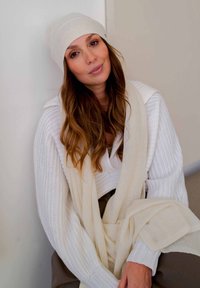 Pull en maille blanc avec des manches surdimensionnées, associé à une écharpe beige et un bonnet assorti. Les cheveux sont ondulés et longs, sur fond neutre.