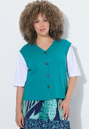 Donna con capelli ricci indossa un gilet verde petrolio con bottoni sopra una maglietta bianca a maniche corte e una gonna fantasia blu-verde, in piedi davanti a uno sfondo bianco.