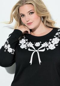 Czarna bluza z białym nadrukiem kwiatowym i wzorem kokardy wzdłuż dekoltu. Posiada długie rękawy i luźny krój. Miękka tekstura tkaniny.