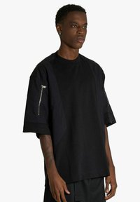 T-shirt nera oversize con maniche corte e dettagli con cerniere, realizzata in tessuto morbido e strutturato, dal design pulito e minimalista.