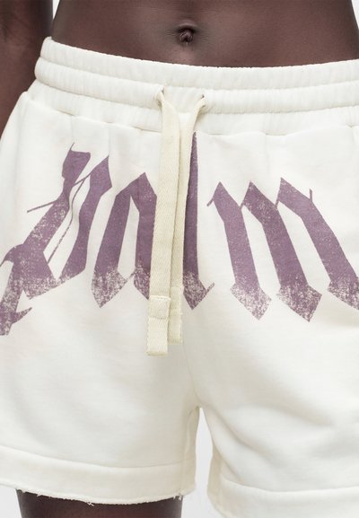 Κοντινό σε μέση ατόμου που φοράει εκρού σορτς με κορδόνι και ξεθωριασμένο σκούρο μωβ γοτθικό κείμενο μπροστά.