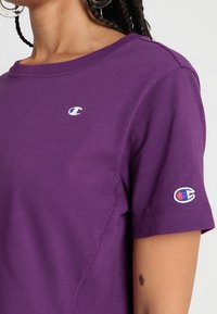 Fialové bavlněné tričko s kulatým výstřihem a krátkými rukávy. Na hrudi a levém rukávu je vyšitý malý logo.