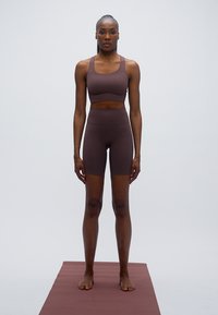 Καφέ αθλητικό σουτιέν και σορτς με ψηλή μέση από ελαστικό ύφασμα, εφαρμοστή σχεδίαση, ομαλή υφή και απλό χρώμα, που στέκεται πάνω σε χαλάκι που ταιριάζει.