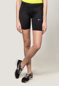 Shorts de sport noirs à la coupe ajustée, texture lisse, avec un logo Nike sur la cuisse gauche. Portés avec des chaussures de sport noires et blanches.