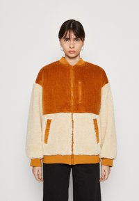 Chaqueta de pana naranja y forro polar crema con cierre frontal, puños acanalados y dos bolsillos delanteros. Presenta un diseño texturizado y con bloques de color.