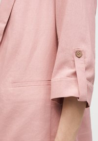 Rukáv svetloružového saka vyhrnutý a zafixovaný drevenou gombíkovou pätkou, detail textúry látky a horizontálneho vrecka.