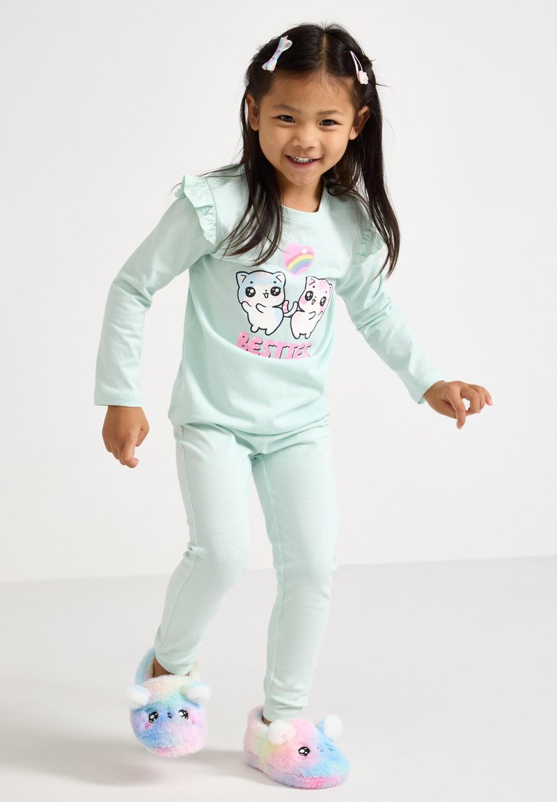 Licht muntgroene langemouwen top met gerimpelde schouders, voorzien van twee cartoonkatten en de tekst "BESTIE". In combinatie met bijpassende broek en kleurrijke pluche slippers.