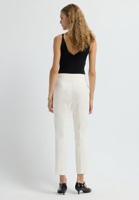 Black sleeveless top with a V-neck, paired with high-waisted white trousers. Model wears shiny black closed-toe shoes. Minimalistic design.