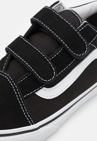 Baskets noires avec coutures blanches, deux bandes velcro noires et une bande blanche incurvée sur le côté, vue rapprochée.