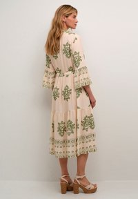 Maxi dress in light beige with green floral patterns, featuring loose bell sleeves, a waist tie, and tiered skirt. Worn with chunky platform heels.