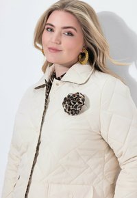 Junge Frau mit blonden Haaren trägt eine gesteppte beige Jacke mit einem Leopardenmuster-Blumenanstecker und goldenen Creolen vor einem weißen Hintergrund.