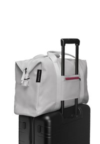 Sac de voyage gris avec double poignée et poche frontale zippée, monté sur une valise noire avec poignée extensible. Texture lisse, design minimaliste.