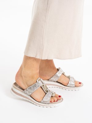 Voeten met rode nagellak die zilveren sandaaltjes met kruisbandjes en witte zolen dragen, gecombineerd met beige geribbelde broekspijpen die net boven de enkels zijn afgewerkt.