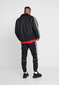 Svart vindjacka med vita ränder längs ärmarna, tillsammans med svarta joggers med en vit rand och svarta träningsskor.