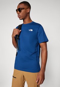 Zila T-krekls, kas izgatavots no kokvilnas, ar mazu baltu logotipu uz krūtīm. Valkāts ar brūnām biksēm un melnu mugursomas siksnu pāri plecam.