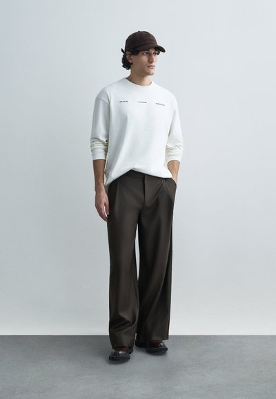 Camisola branca de manga comprida com texto preto, combinada com calças castanhas largas. Inclui um boné castanho e sapatos pretos. Superfícies texturizadas.