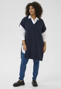 Pulover tip poncho de culoare bleumarin, tricotat, cu guler în V și fante laterale, purtat peste o cămașă albă cu butoni și asortat cu blugi blue și pantofi negri.