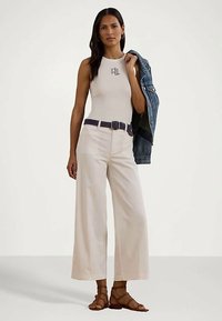 Mulher vestindo uma blusa branca sem mangas com logotipo, calças largas creme com cinto, sandálias castanhas, segurando uma jaqueta de ganga sobre um ombro.