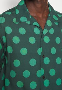 Chemise vert foncé avec de grands pois verts lumineux, portée par une personne à la peau foncée, montrant le col et le haut de la poitrine.
